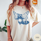 Comfort Colors tee in Ivory featuring whimsical graphic style of a raccoon with large detailed butterfly wings