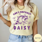 Comfort Colors tee in Butter featuring whimsical raccoon graphic with daisy flowers and God's Oopsiest Daisy text