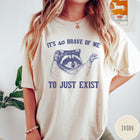 Comfort Colors tee in Ivory featuring illustrative raccoon graphic with text It's So Brave Of Me To Just Exist