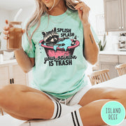 Comfort Colors tee in Island Reef featuring illustrative graphic of a raccoon sitting in a pink bathtub