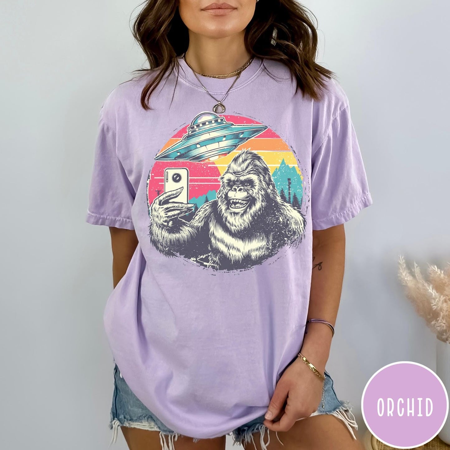 Comfort Colors tee in Orchid featuring retro Bigfoot selfie graphic with a UFO and sunset mountain background