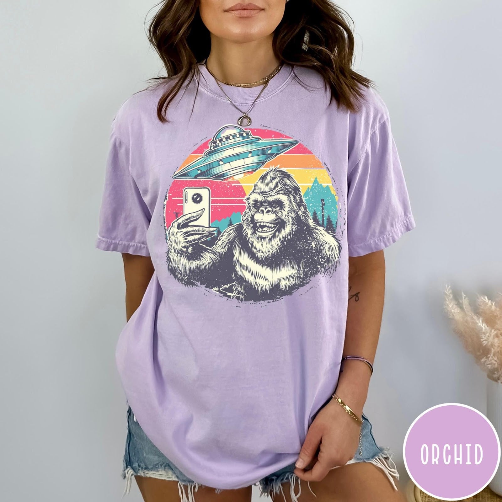 Comfort Colors tee in Orchid featuring retro Bigfoot selfie graphic with a UFO and sunset mountain background