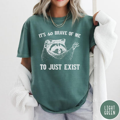 Comfort Colors tee in Light Green featuring illustrative raccoon graphic with text It's So Brave Of Me To Just Exist