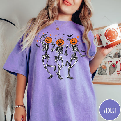 Comfort Colors tee in Violet featuring dancing skeleton graphic with jack-o'-lantern heads and small orange stars