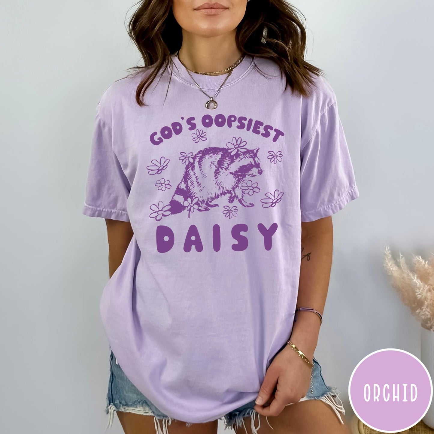 Comfort Colors tee in Orchid featuring whimsical raccoon graphic with daisy flowers and God's Oopsiest Daisy text