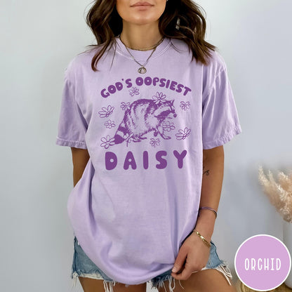 Comfort Colors tee in Orchid featuring whimsical raccoon graphic with daisy flowers and God's Oopsiest Daisy text