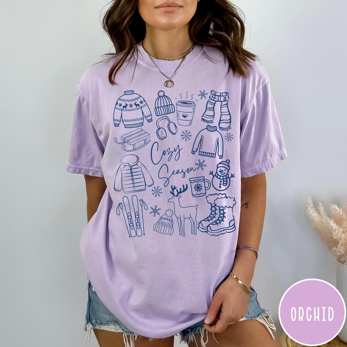 Comfort Colors tee in Orchid featuring winter activities graphic with sweaters, sled, snowman, and seasonal items.