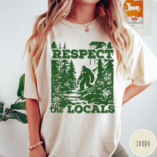 Comfort Colors tee in Ivory featuring vintage style graphic of Bigfoot walking through a forest by a river