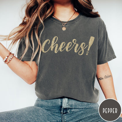 Comfort Colors tee in Pepper featuring New Year celebration graphic with gold glitter cheers text and champagne bottle