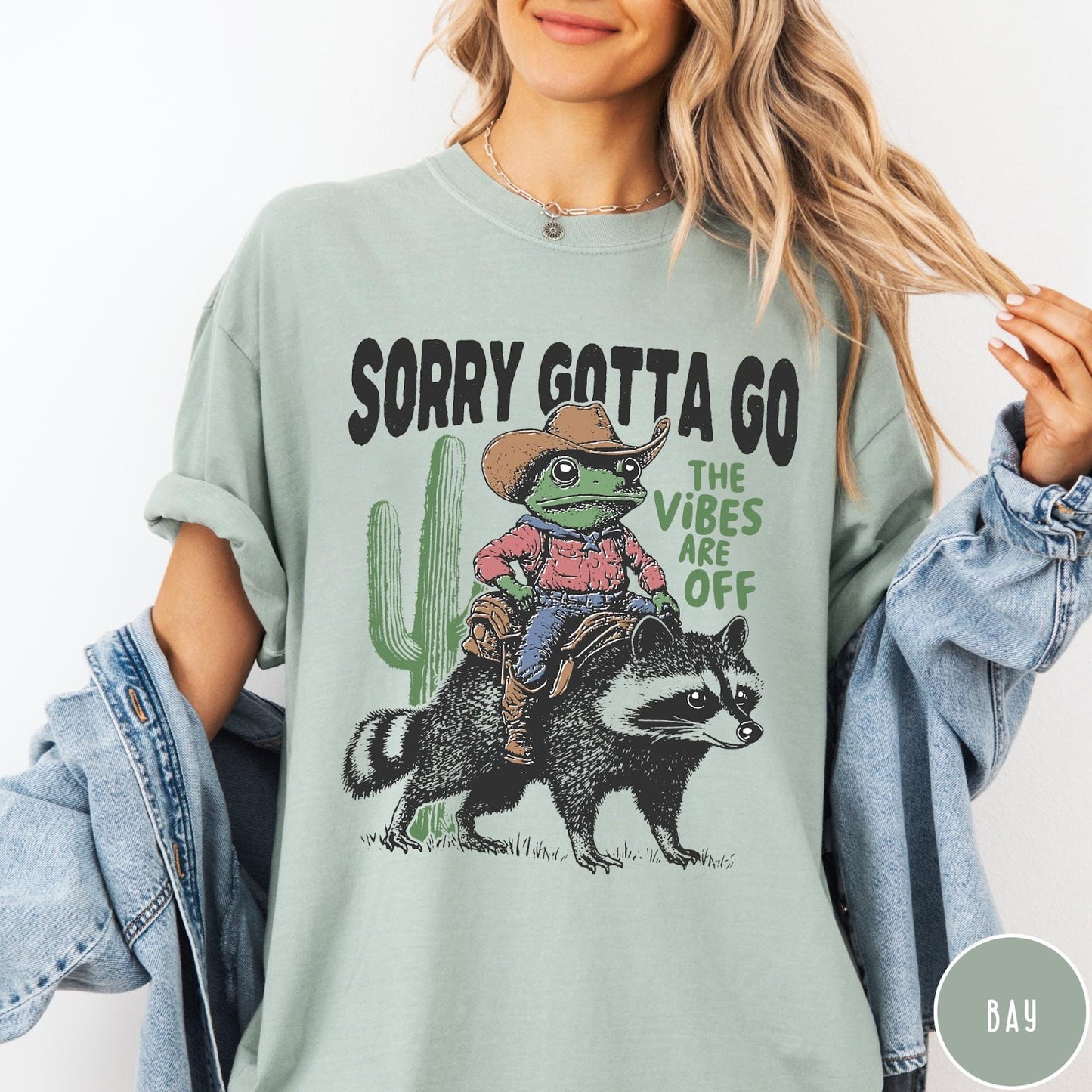 Comfort Colors tee in Bay featuring western graphic of a cowboy frog riding a raccoon with cactus
