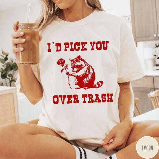 Comfort Colors tee in Ivory featuring a whimsical graphic of a raccoon holding a red rose.