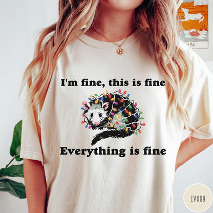 Comfort Colors tee in Ivory featuring graphic illustration of an opossum tangled in colorful Christmas lights