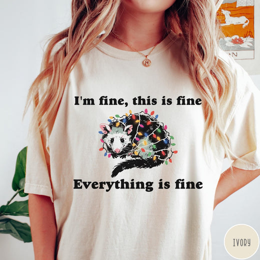 Comfort Colors tee in Ivory featuring graphic illustration of an opossum tangled in colorful Christmas lights