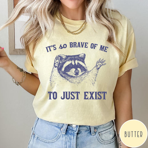Comfort Colors tee in Butter featuring illustrative raccoon graphic with text It's So Brave Of Me To Just Exist