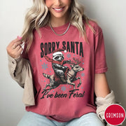 Comfort Colors tee in Crimson featuring festive graphic of a raccoon wearing a Santa hat riding a reindeer