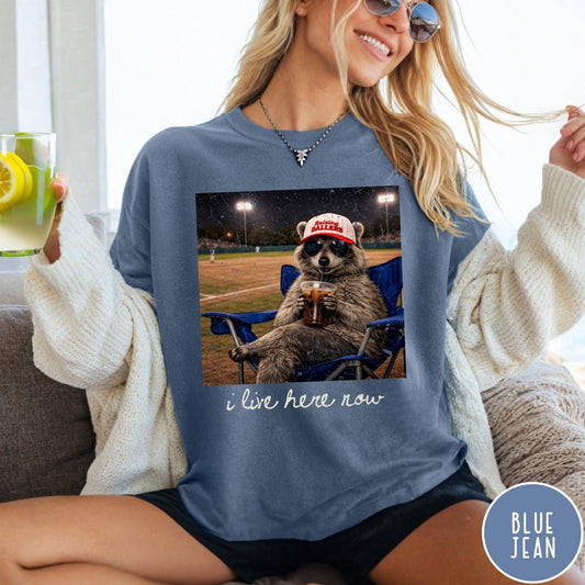 Comfort Colors tee in Blue Jean featuring graphic style raccoon wearing sunglasses and baseball cap at a stadium
