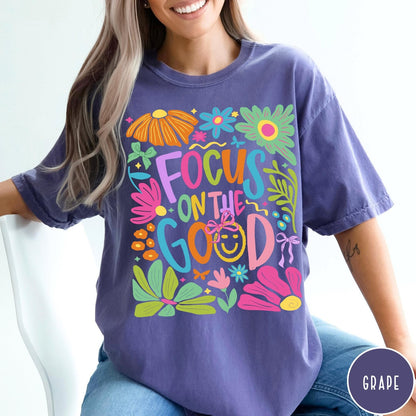 Comfort Colors tee in Grape featuring positivity typography and floral collage graphic with a smiley face.