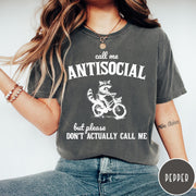 Comfort Colors tee in Pepper featuring graphic style illustration of a raccoon riding a bicycle
