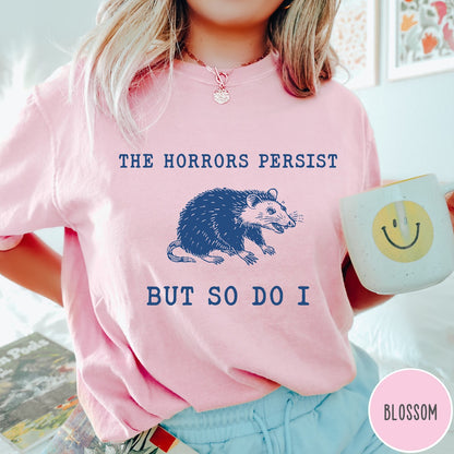 Comfort Colors tee in Blossom featuring illustrative style opossum graphic with text The Horrors Persist But So Do I