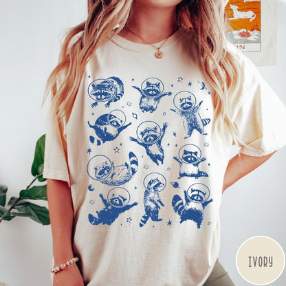 Comfort Colors tee in Ivory featuring whimsical space design with multiple raccoons wearing glass astronaut helmets and stars
