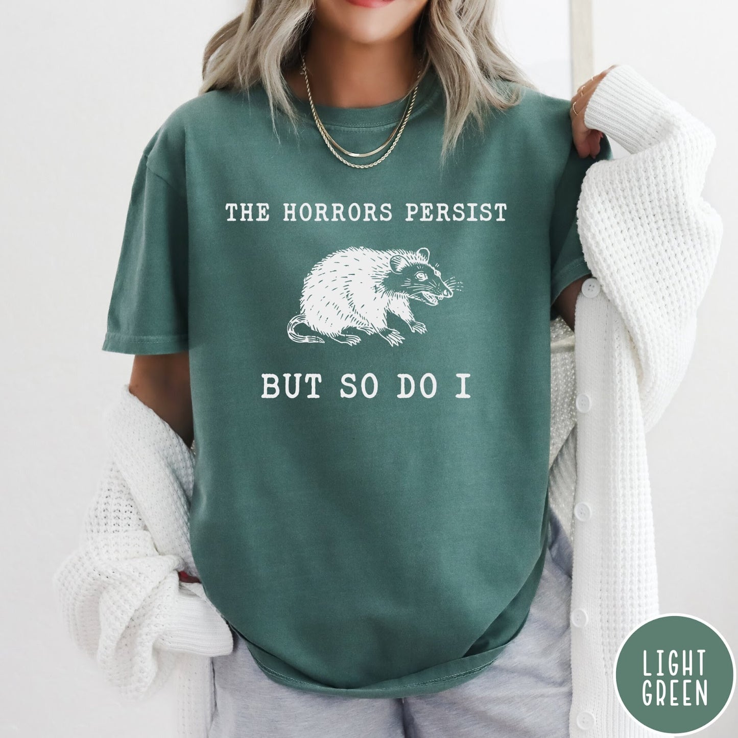 Comfort Colors tee in Light Green featuring illustrative style opossum graphic with text The Horrors Persist But So Do I
