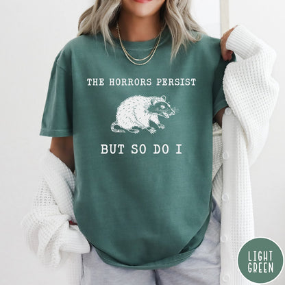 Comfort Colors tee in Light Green featuring illustrative style opossum graphic with text The Horrors Persist But So Do I