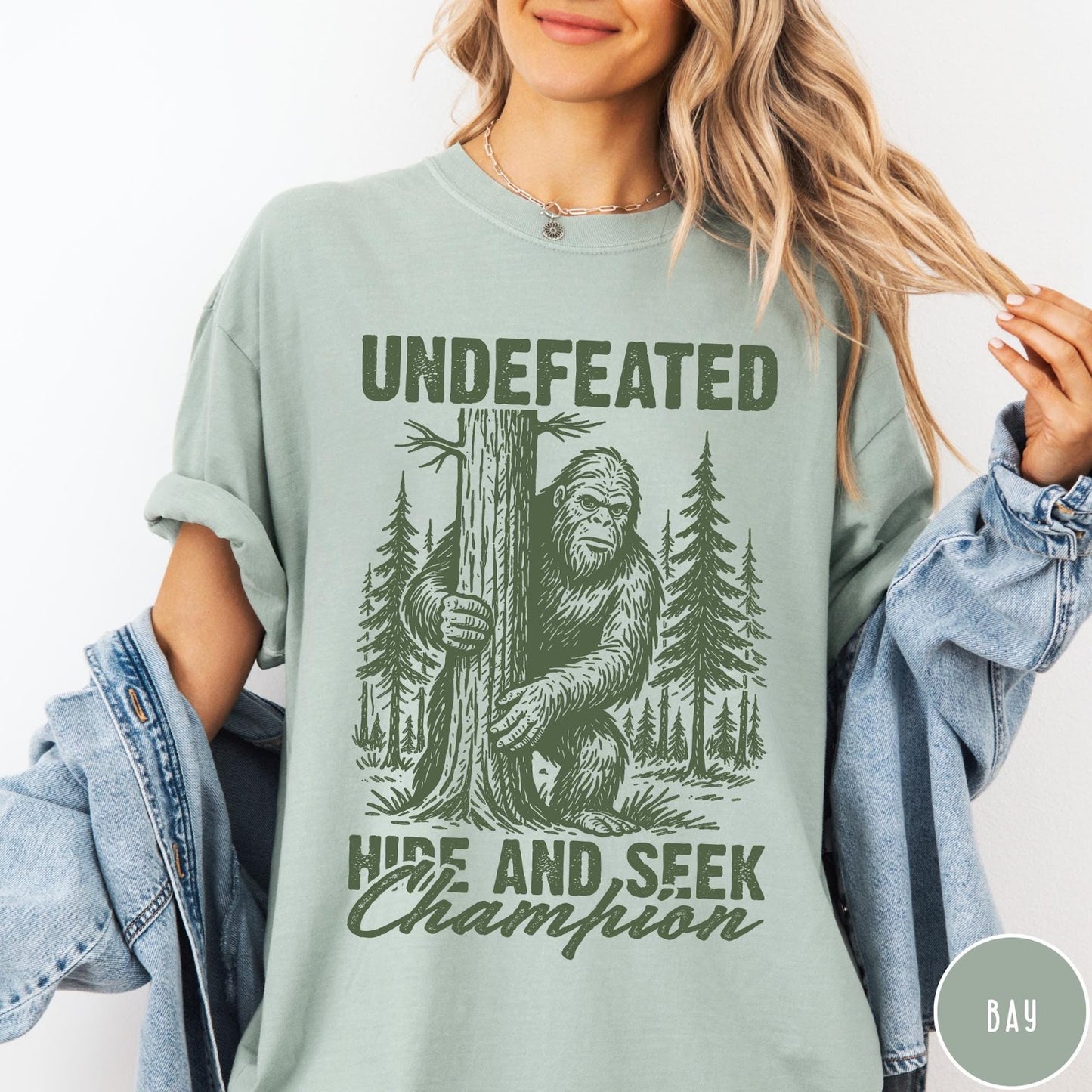Comfort Colors tee in Bay featuring vintage style Bigfoot graphic hiding behind a tree in the forest