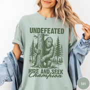 Comfort Colors tee in Bay featuring vintage style Bigfoot graphic hiding behind a tree in the forest