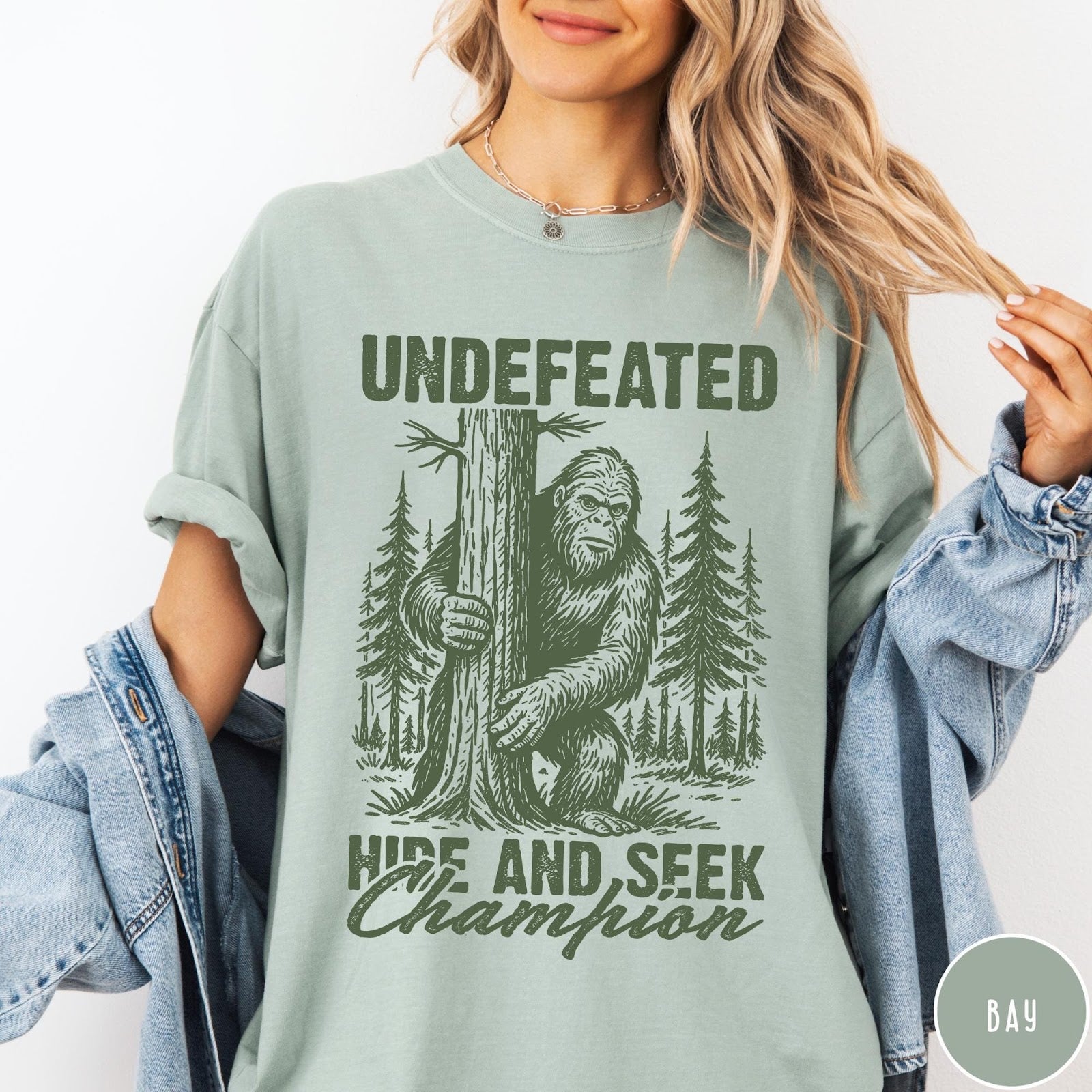 Comfort Colors tee in Bay featuring vintage style Bigfoot graphic hiding behind a tree in the forest