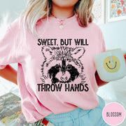 Comfort Colors tee in Blossom featuring graphic style raccoon illustration with text Sweet But Will Throw Hands