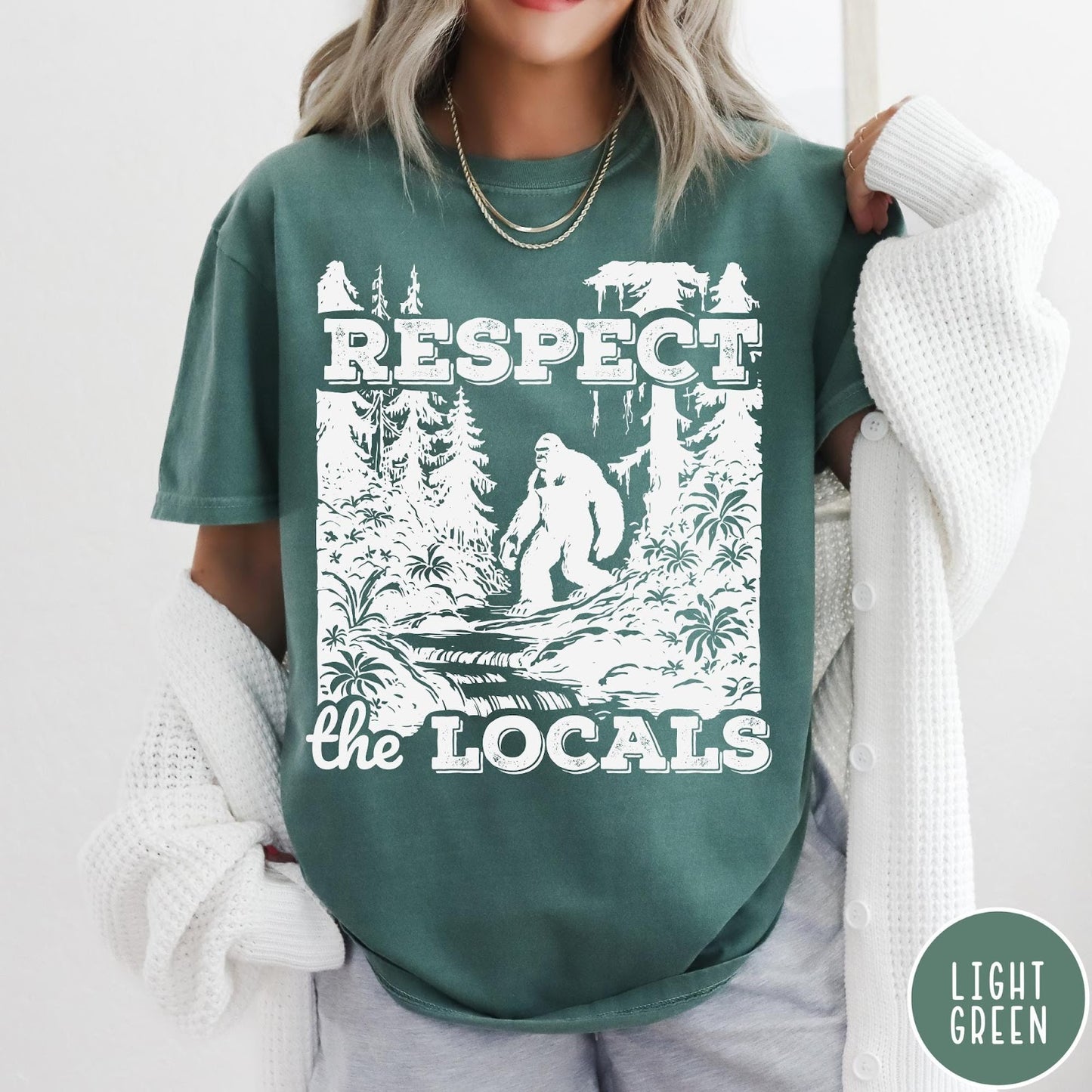 Comfort Colors tee in Light Green featuring vintage style graphic of Bigfoot walking through a forest by a river