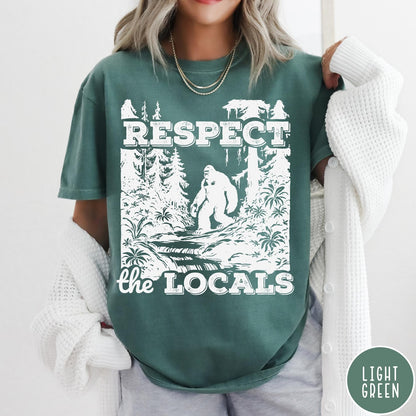 Comfort Colors tee in Light Green featuring vintage style graphic of Bigfoot walking through a forest by a river