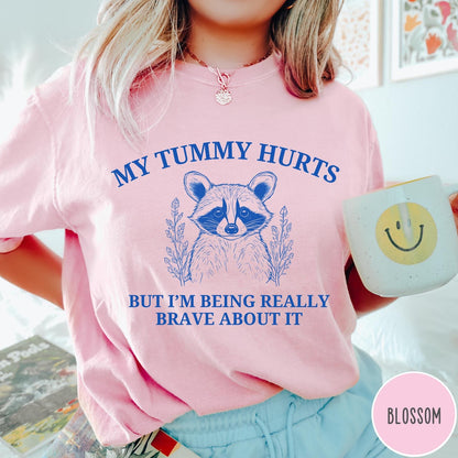 Comfort Colors tee in Blossom featuring line art style raccoon graphic with My Tummy Hurts text design