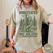 Comfort Colors tee in Khaki featuring vintage style Bigfoot graphic hiding behind a tree in the forest