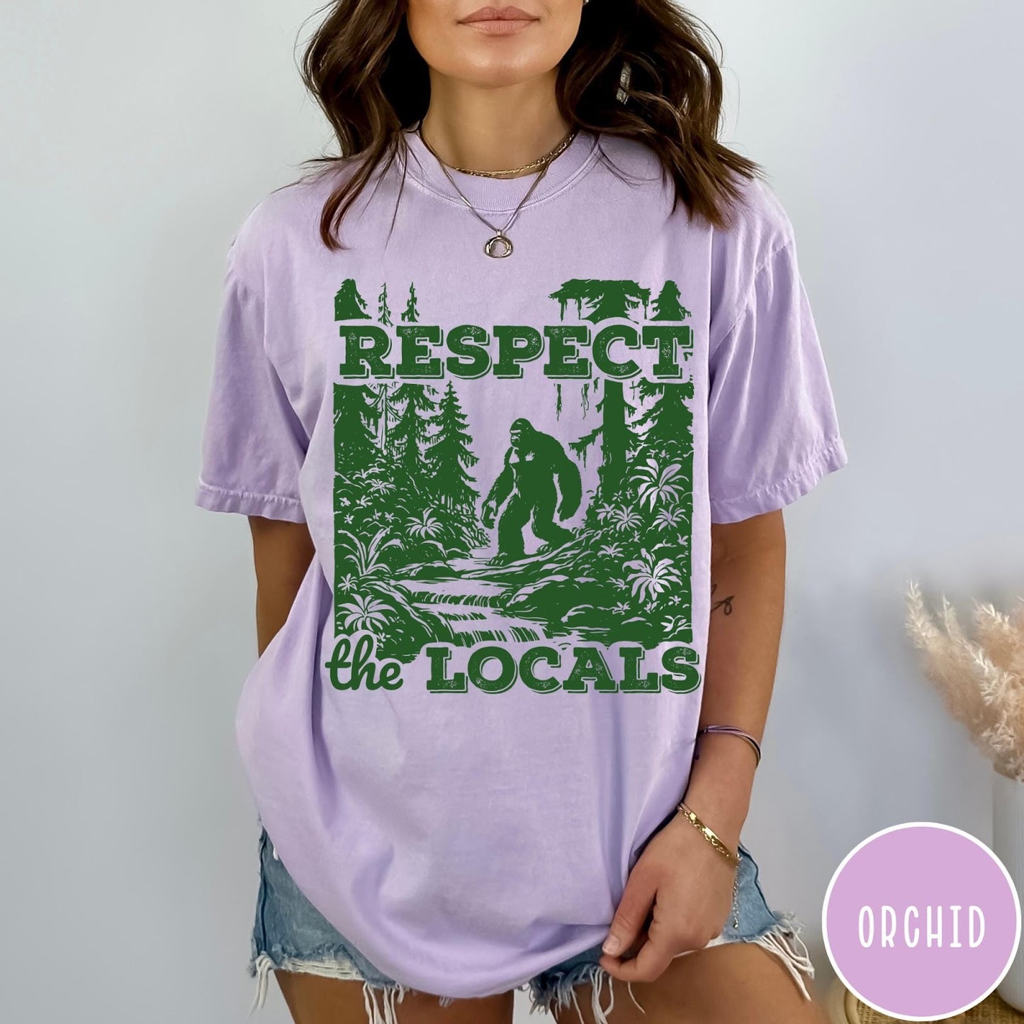 Comfort Colors tee in Orchid featuring vintage style graphic of Bigfoot walking through a forest by a river