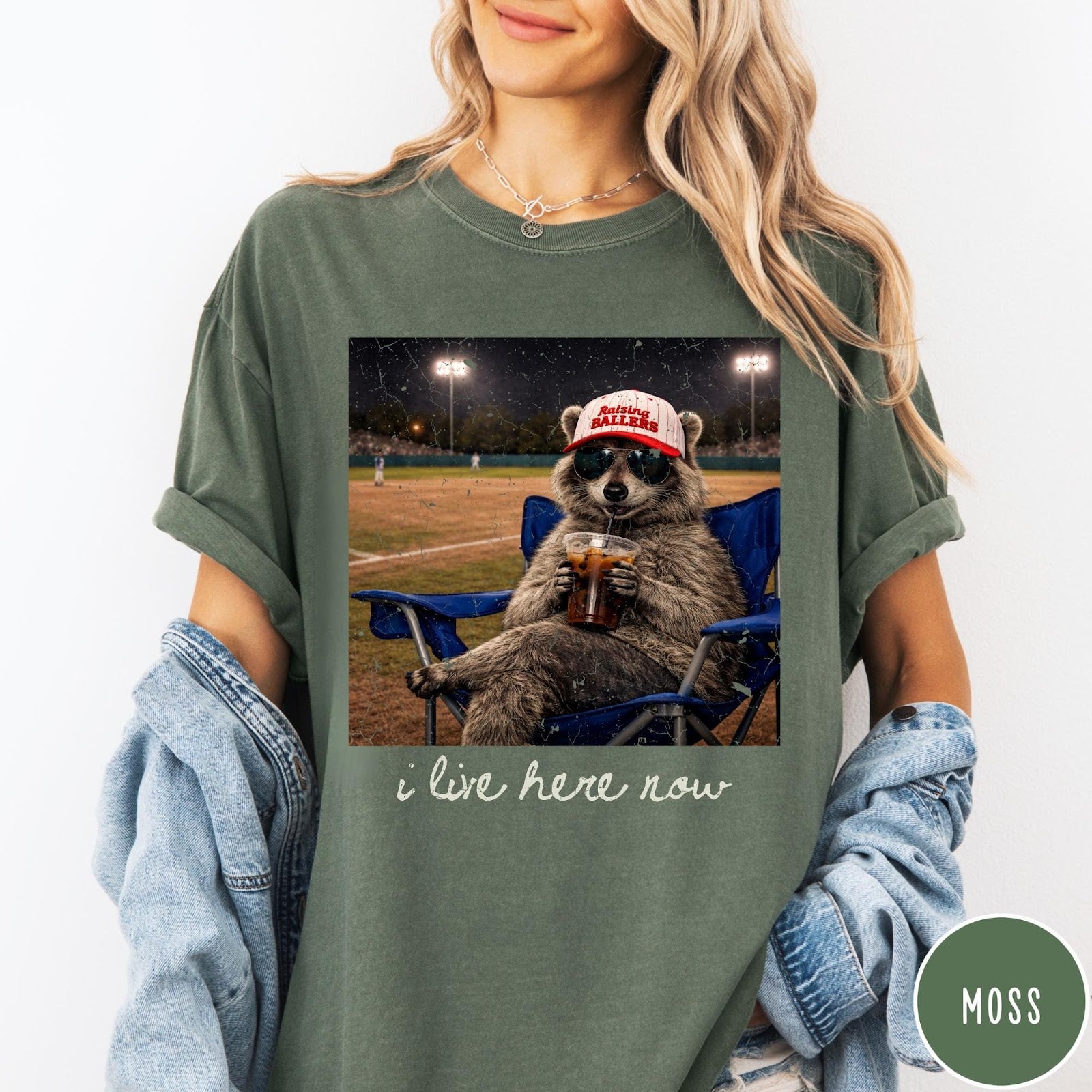 Comfort Colors tee in Moss featuring graphic raccoon wearing sunglasses and baseball cap sitting in camping chair