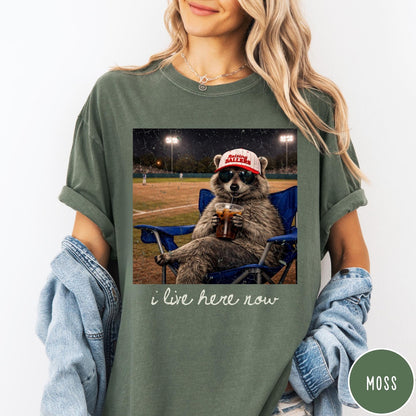 Comfort Colors tee in Moss featuring graphic raccoon wearing sunglasses and baseball cap sitting in camping chair