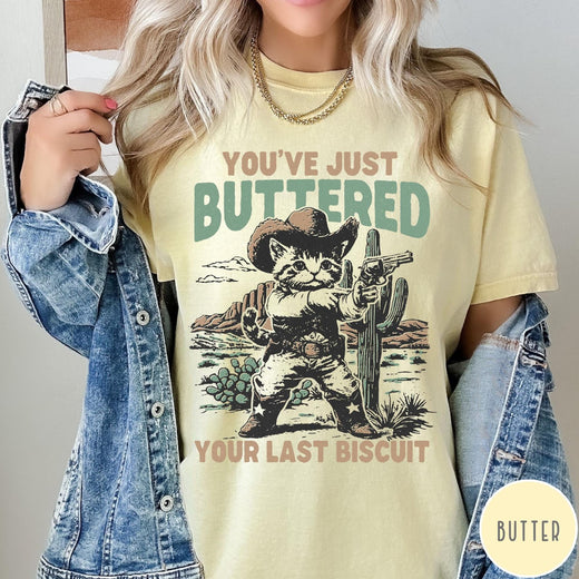 Comfort Colors tee in Butter featuring western graphic design of a cat wearing a cowboy hat and gun.