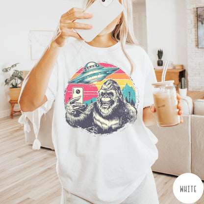 Comfort Colors tee in White featuring retro Bigfoot selfie graphic with a UFO and sunset mountain background