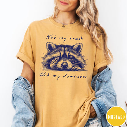 Comfort Colors tee in Mustard featuring graphic illustration of a winking raccoon with text Not My Trash