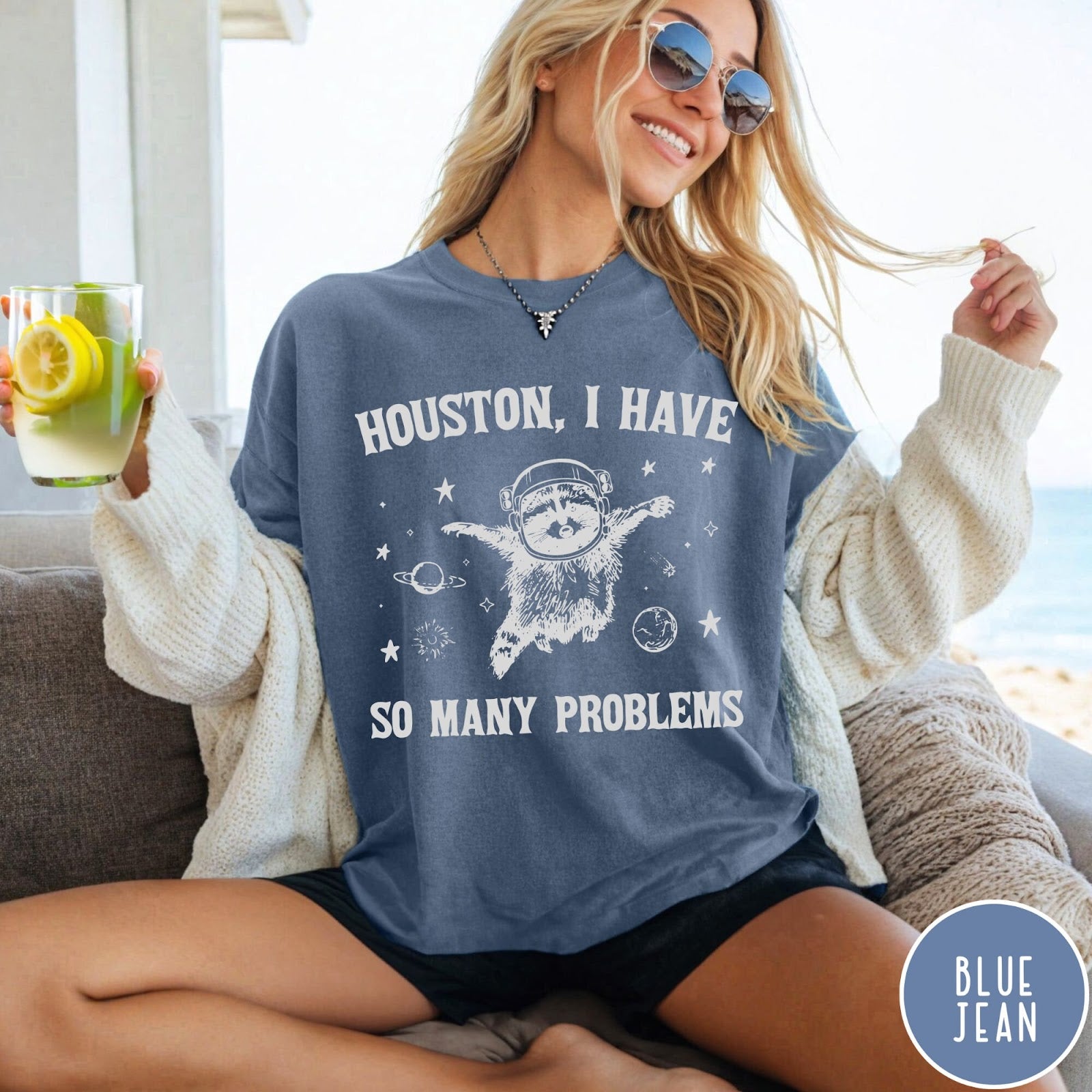 Comfort Colors tee in Blue Jean featuring space style graphic of a raccoon wearing an astronaut helmet floating