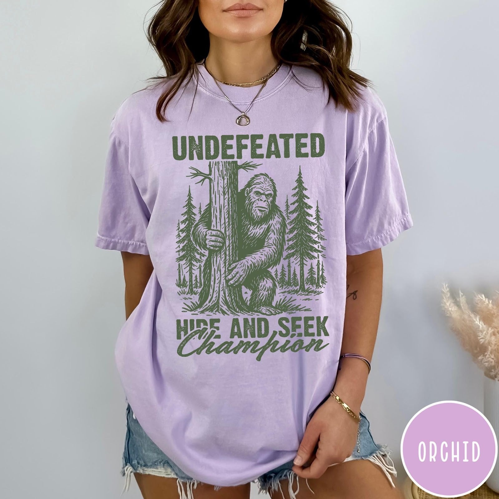 Comfort Colors tee in Orchid featuring vintage style graphic of Bigfoot hiding behind a large pine tree