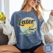 Comfort Colors tee in Blue Jean featuring vintage style graphic of an alligator emerging from water with sunset background