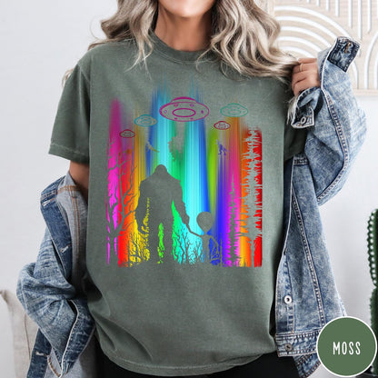 Comfort Colors tee in Moss featuring a vibrant graphic of Bigfoot and an alien holding hands in forest
