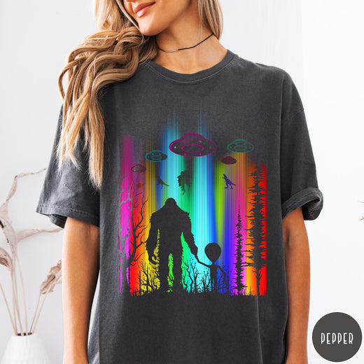 Comfort Colors tee in Pepper featuring a vibrant graphic of Bigfoot and an alien holding hands in forest