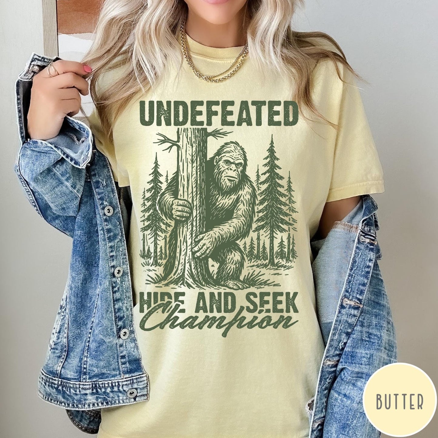 Comfort Colors tee in Butter featuring vintage style graphic of Bigfoot hiding behind a large pine tree