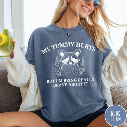 Comfort Colors tee in Blue Jean featuring line art style raccoon graphic with My Tummy Hurts text design