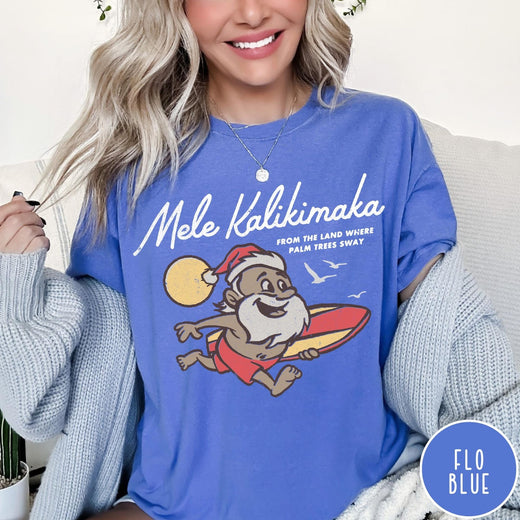 Comfort Colors tee in Flo Blue featuring vintage style graphic of a surfing Santa Claus on vacation