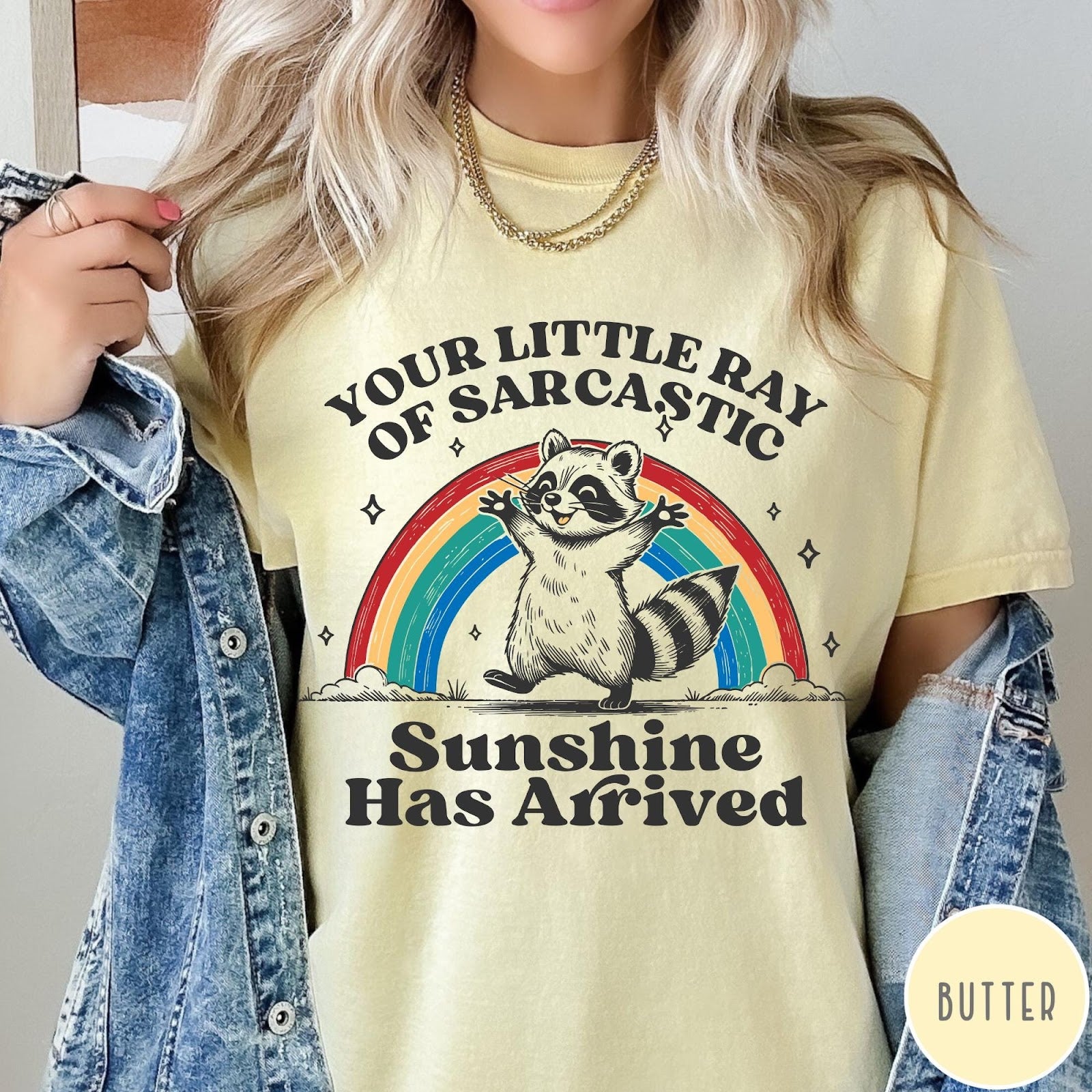 Comfort Colors tee in Butter featuring vintage style graphic of a happy raccoon standing before a colorful rainbow