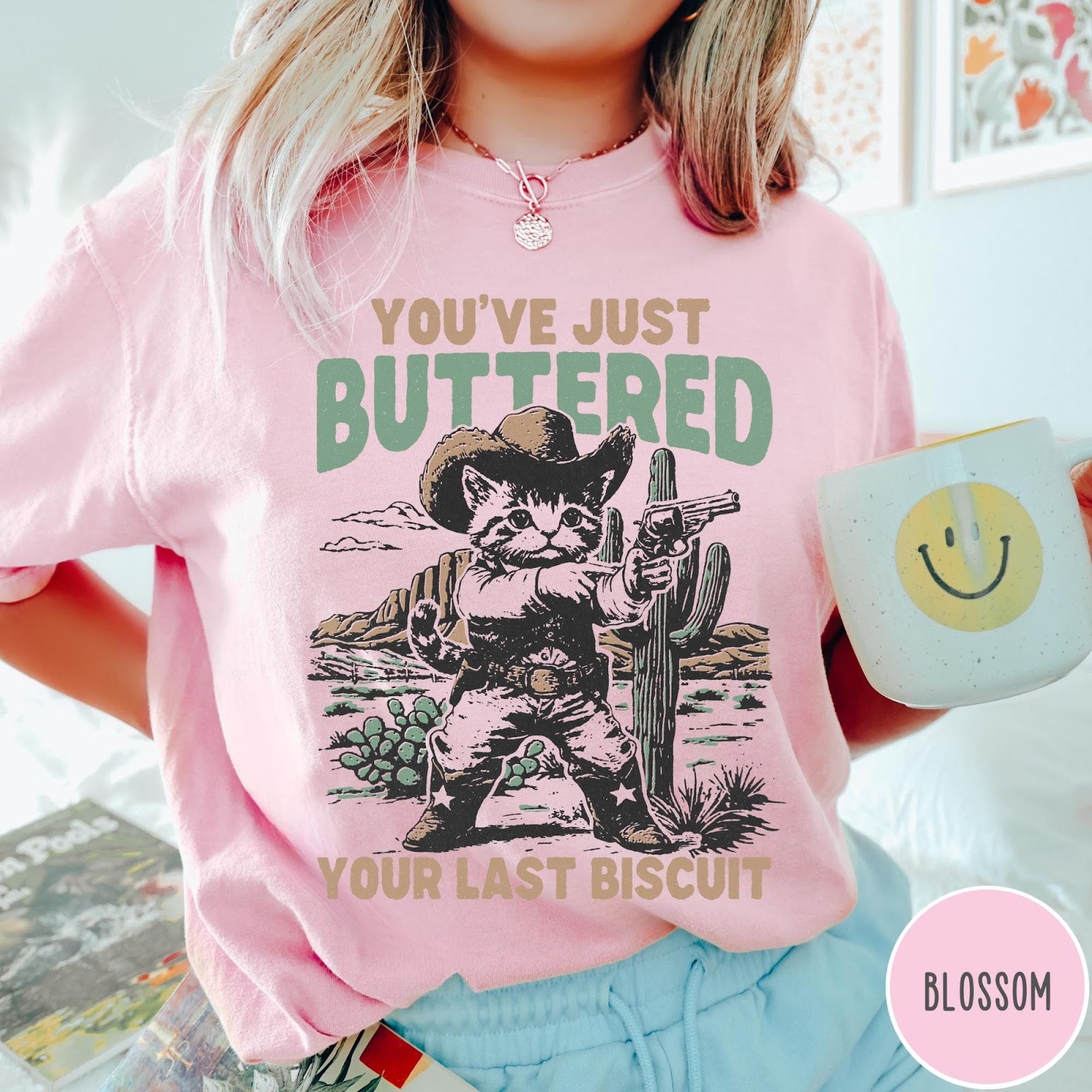 Comfort Colors tee in Blossom featuring western graphic design of a cat wearing a cowboy hat and gun.